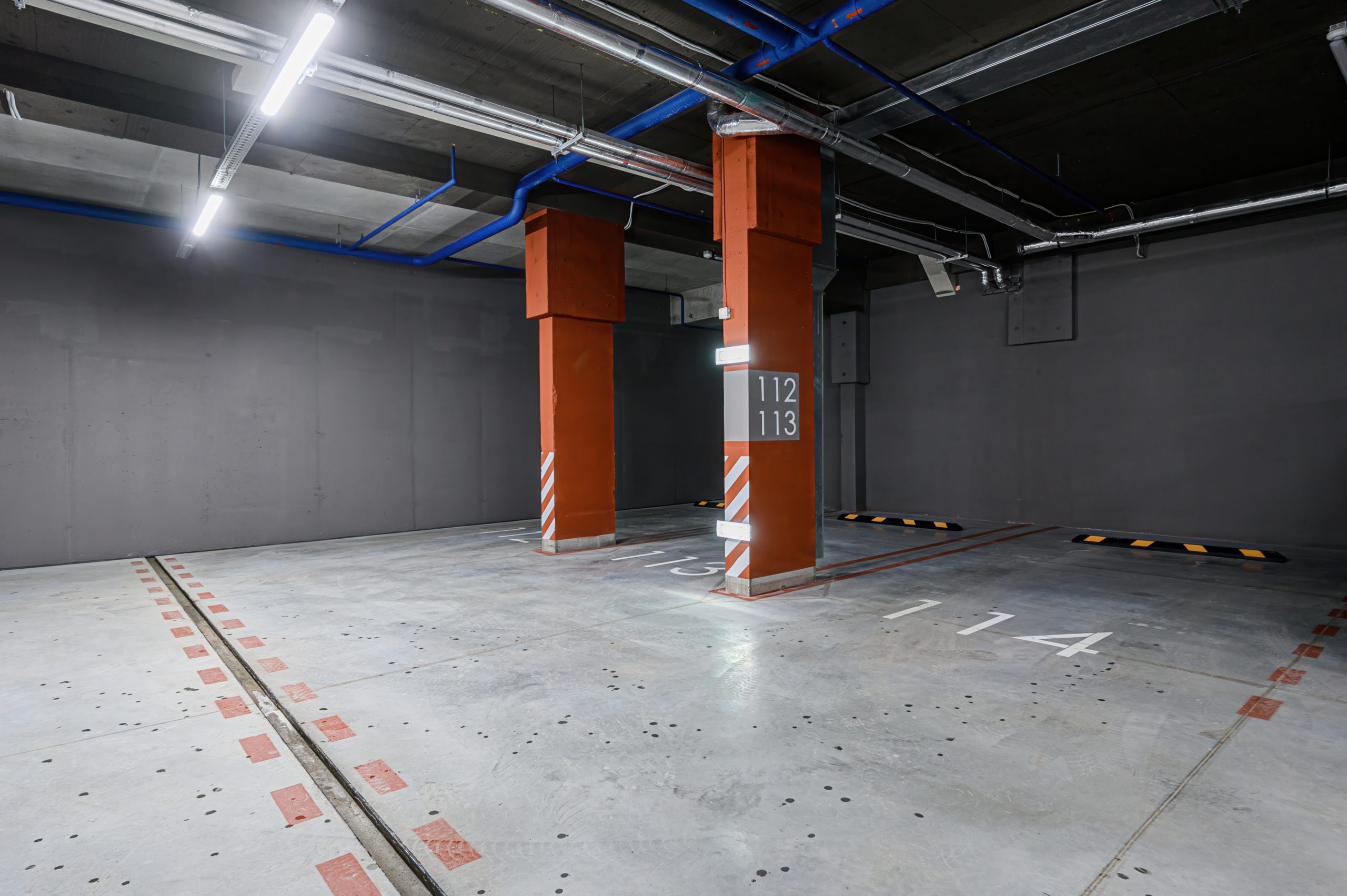 Indoor parking with orange columns, marked spaces, and visible ceiling pipes. Concrete floor and gray walls